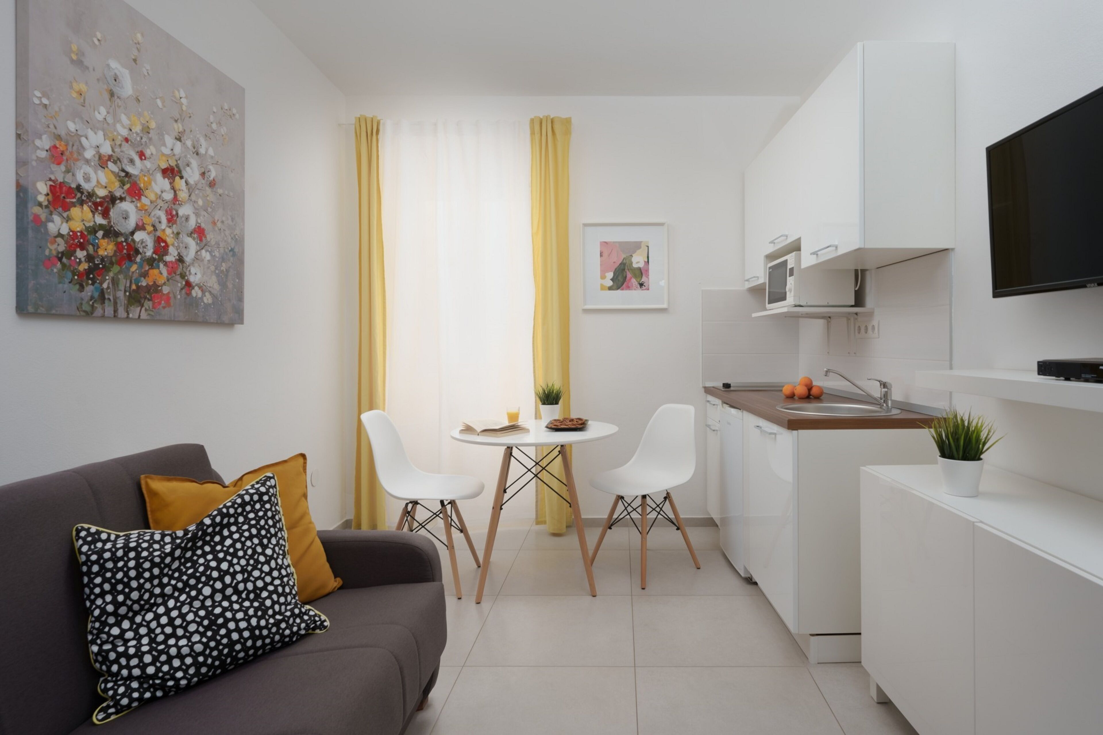 Une cuisine moderne avec des meubles blancs, un four à micro-ondes et un évier. On y trouve un coin repas avec une table et des chaises, ainsi qu’un canapé avec des coussins.