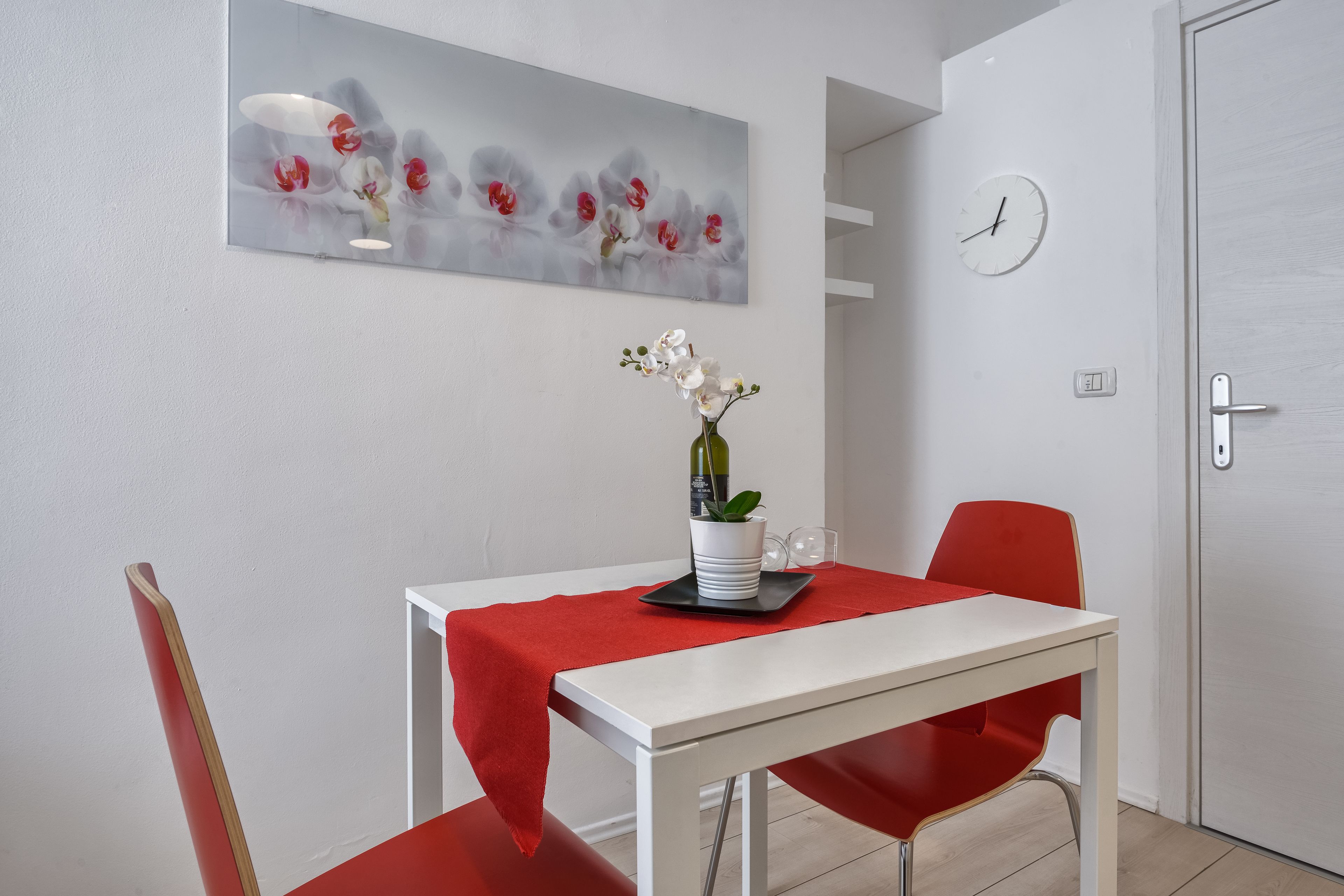 A dining area with a white table, red chairs, a red tablecloth, a vase with flowers, and a clock on the wall.