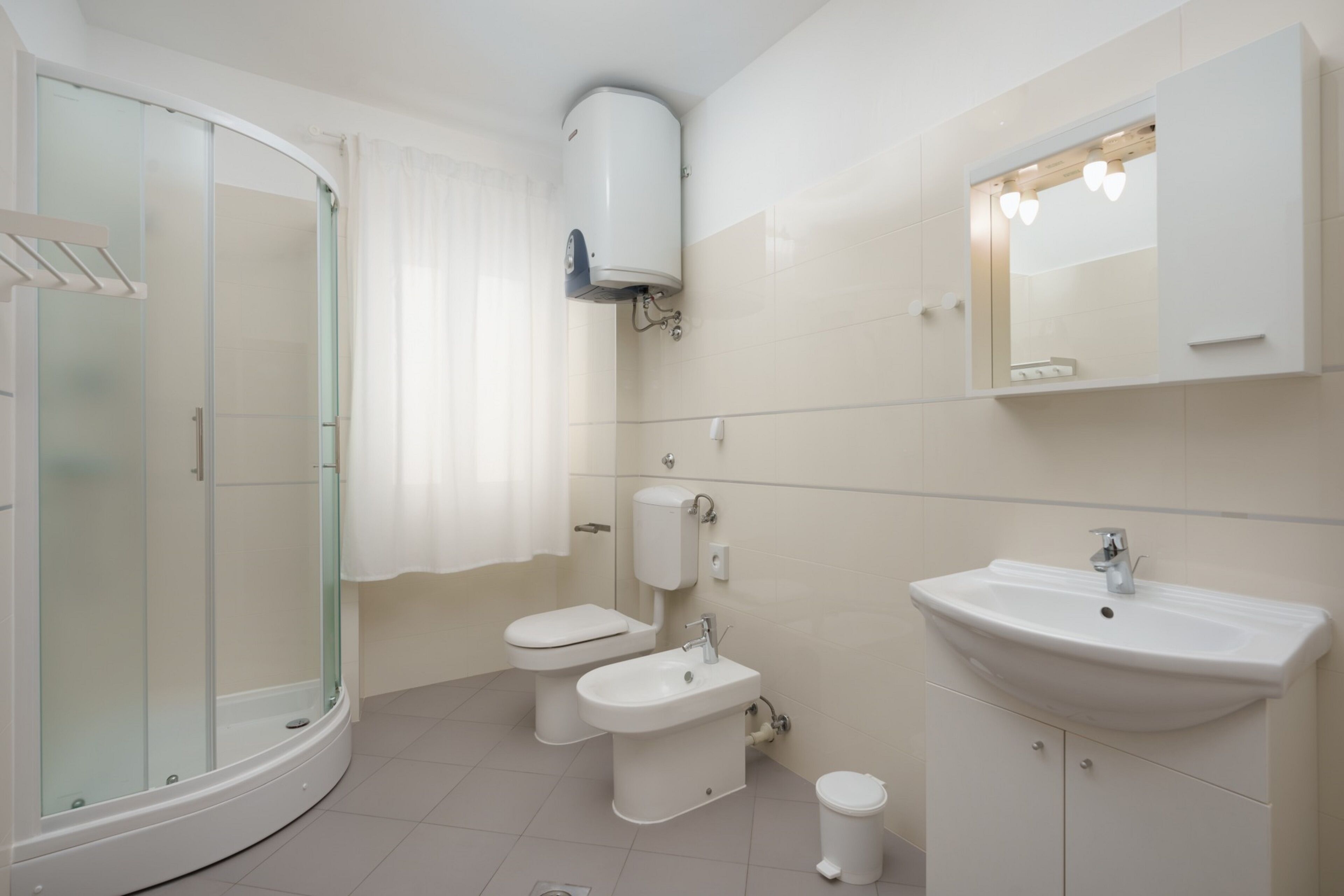 A modern bathroom with a glass shower enclosure, two toilets, a sink, and a mirror with lights.