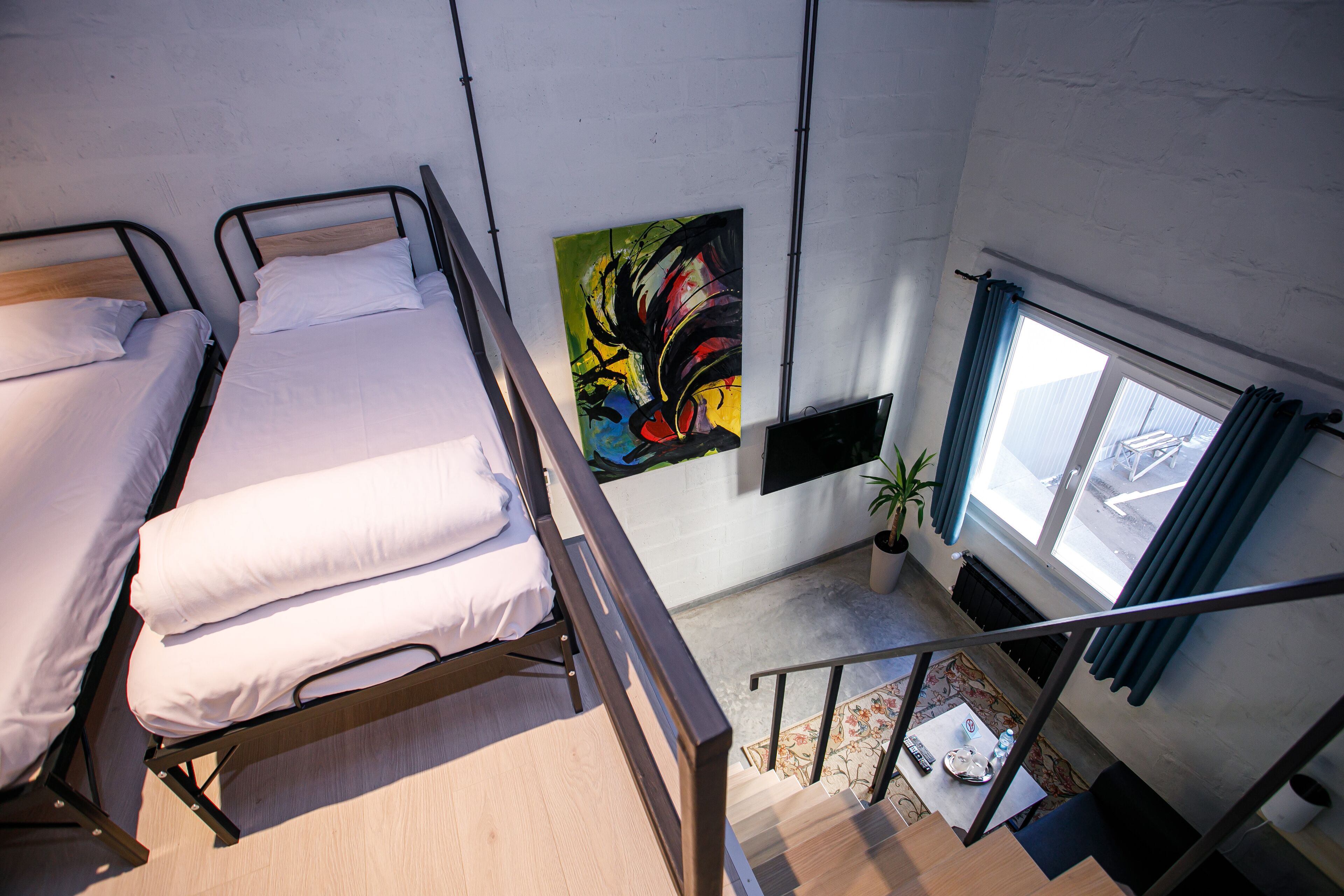 Une chambre de dortoir avec deux lits, un tableau abstrait aux couleurs vives, une fenêtre avec des rideaux et un escalier.