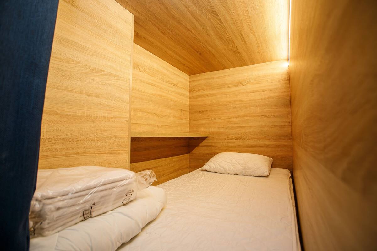A small, compact room with wooden walls and a sloped ceiling. There is a single bed with white bedding and a pillow.