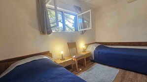 A room with two beds, a window with curtains, a nightstand with a lamp, and a chair.