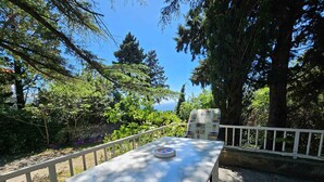 A table with a map and a chair, set on a deck with a white railing, surrounded by trees and a view of the sea.