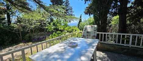 A table with a map and a chair, set on a deck with a white railing, surrounded by trees and a view of the sea.
