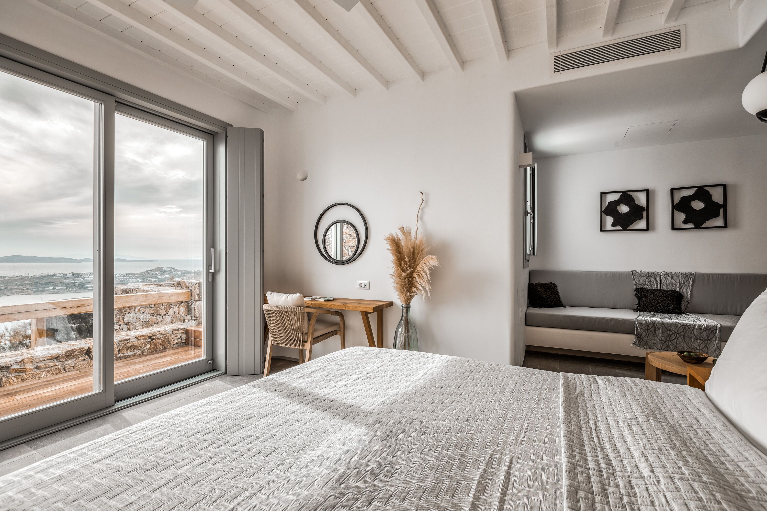 A modern bedroom with a large bed, a desk, and a sofa. There is a view of the city through large windows.
