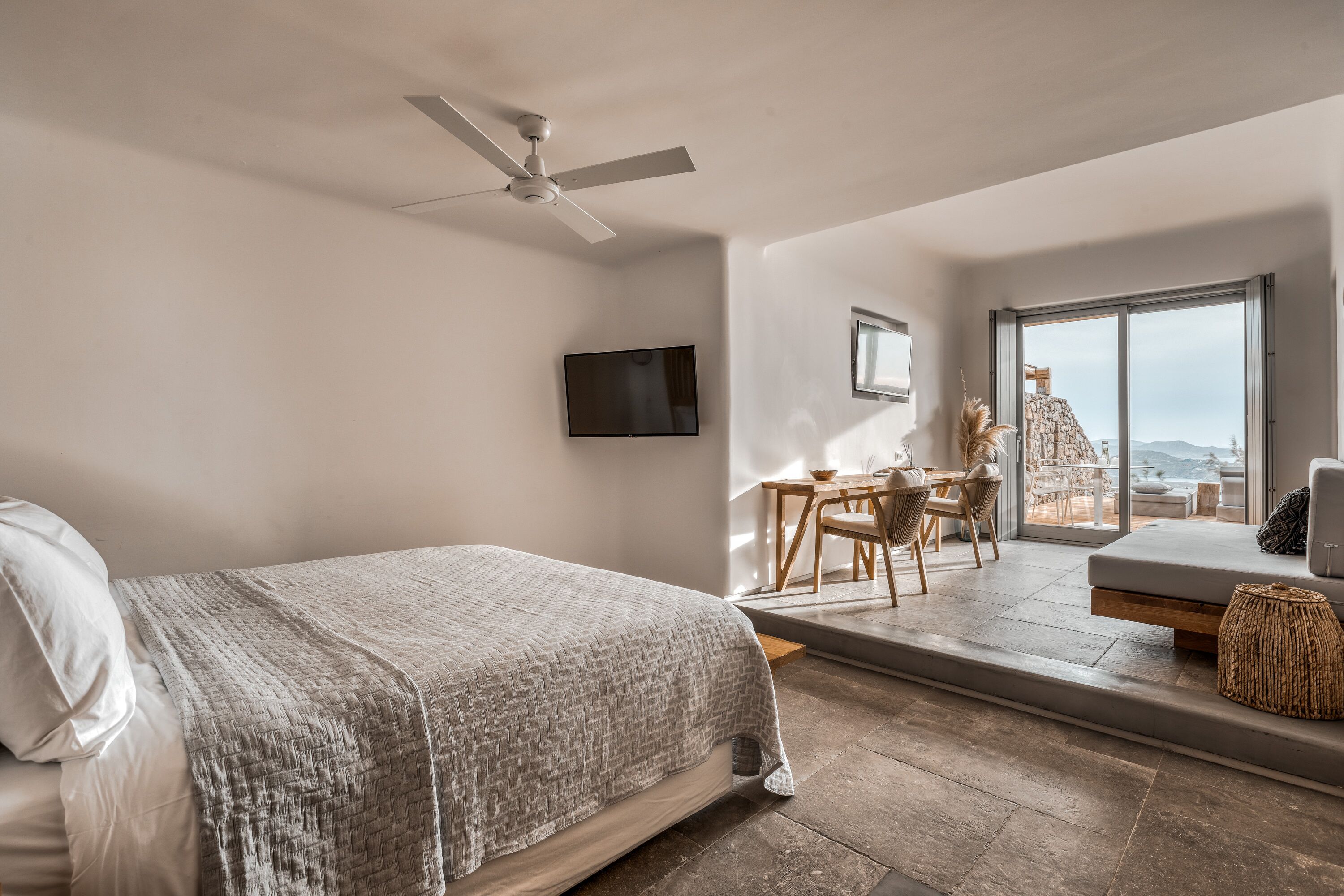 Une chambre avec un lit, un ventilateur de plafond, une télévision, un balcon avec une table et des chaises, et une vue sur l’extérieur.