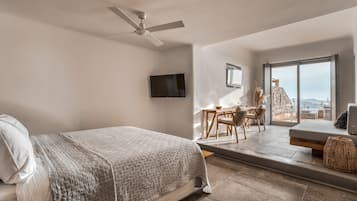 Une chambre avec un lit, un ventilateur de plafond, une télévision, un balcon avec une table et des chaises, et une vue sur l’extérieur.