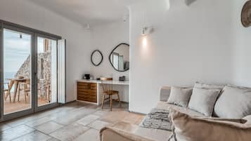 A modern living room with a sofa, a dining table with chairs, a desk with a chair, and a large window with a view of the sea.