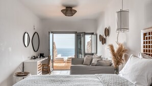 A modern bedroom with a large bed, a desk, and a sofa. There is a view of the ocean through the open door. - Alissachni Mykonos (Mykonos)