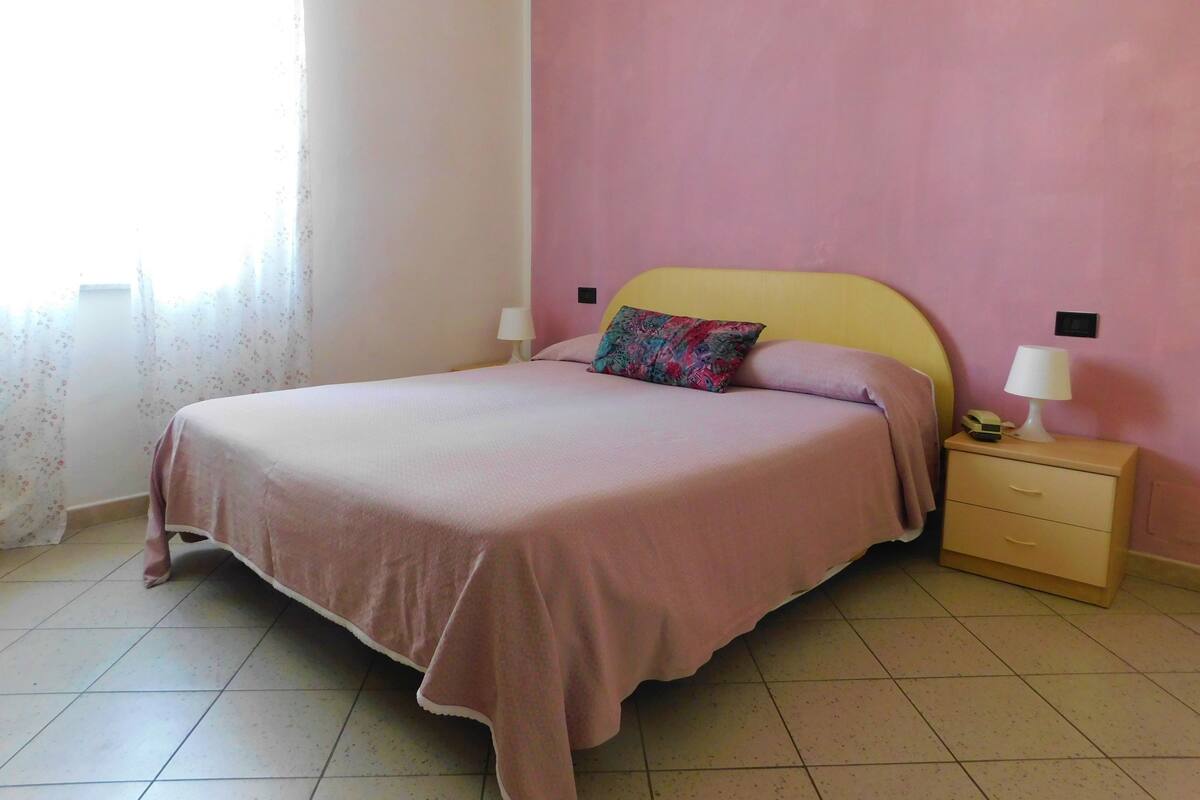 Um quarto com cama, mesa de cabeceira, abajur e relógio de parede.