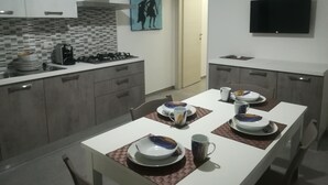 A modern kitchen with grey cabinets, a gas stove, and a microwave. There is a dining table set with plates, bowls, and a coffee maker. A painting hangs on the wall, and a TV is mounted above the counter.