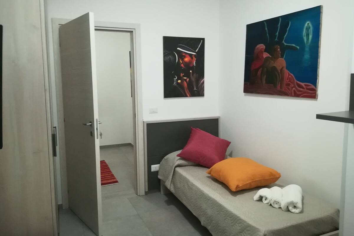 A bedroom with a bed, a red and orange pillow, and two rolled towels. There are two paintings on the wall.