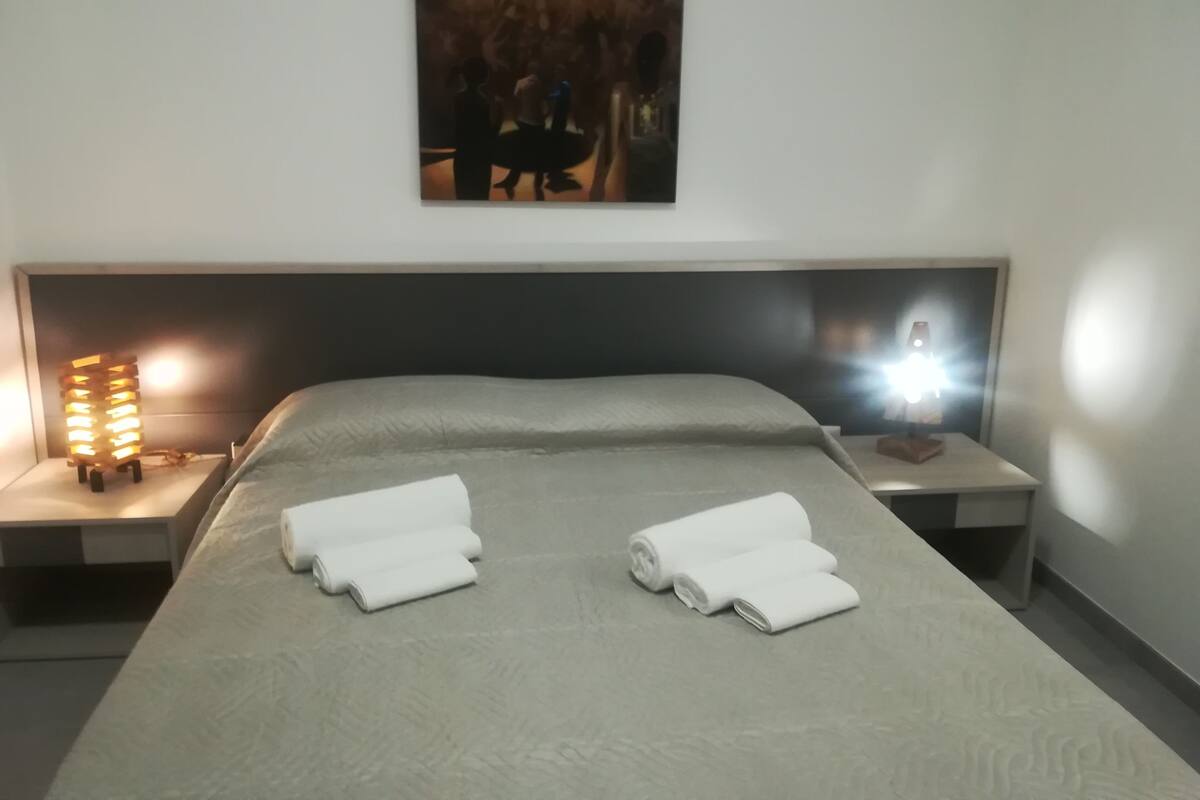 A neatly made bed with two rolled towels, a painting above the bed, and two bedside lamps.