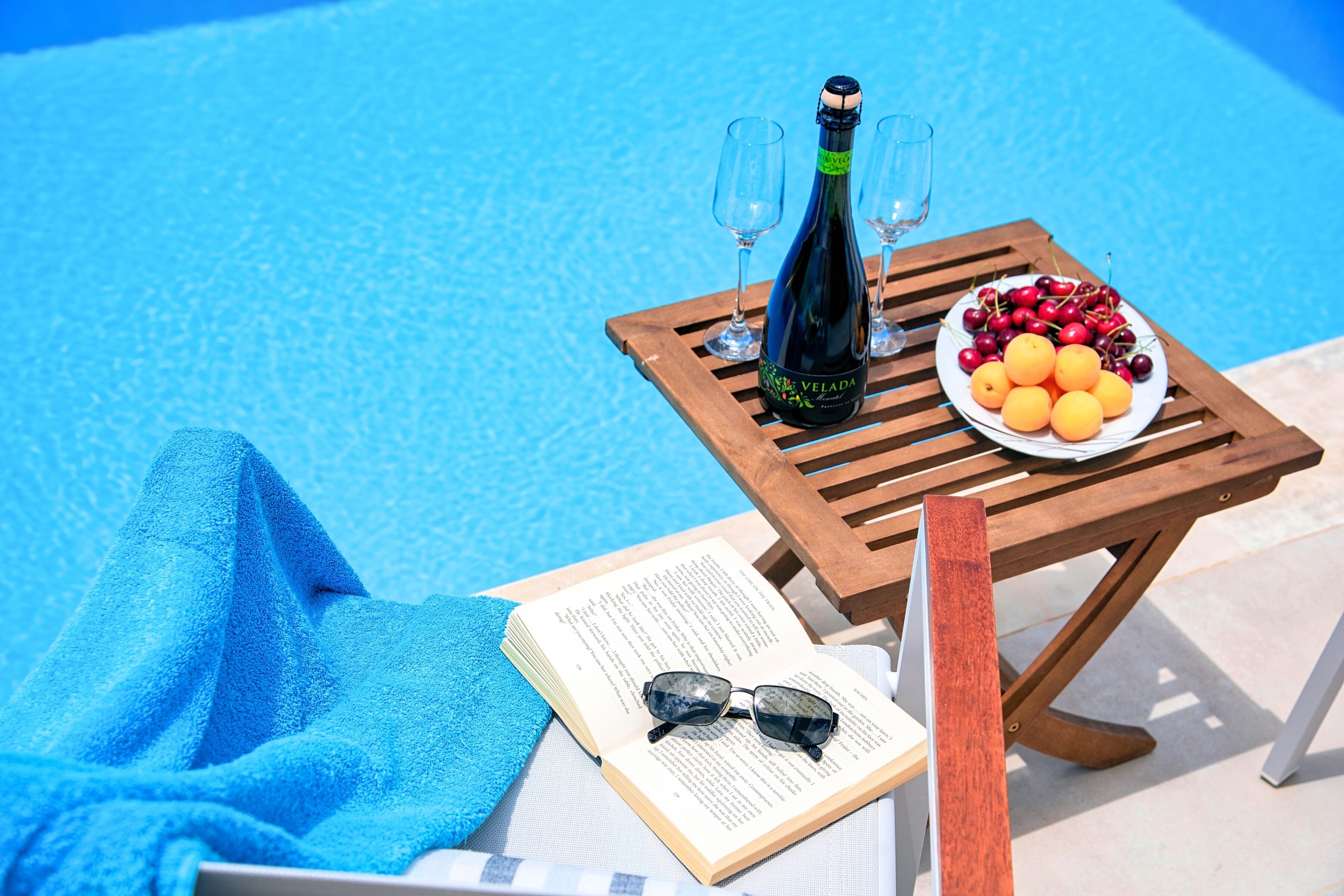 A poolside setting with a wooden table, a bottle of champagne, glasses, a plate of fruit, a towel, and a book.