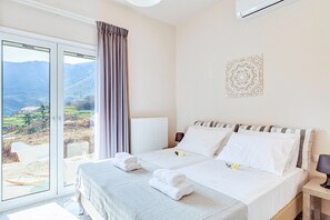 A hotel room with a bed, a bedside table, a lamp, a decorative wall piece, and a view of mountains through a large window. - Villa di Giorgio Heated Pool (Apokoronas)