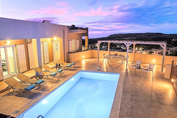 A rooftop pool area with lounge chairs, a pergola, and a view of the city at dusk. - Villa di Giorgio Heated Pool (Apokoronas)