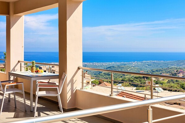 A balcony with a table and chairs, overlooking a scenic view of the sea and a hilly landscape.