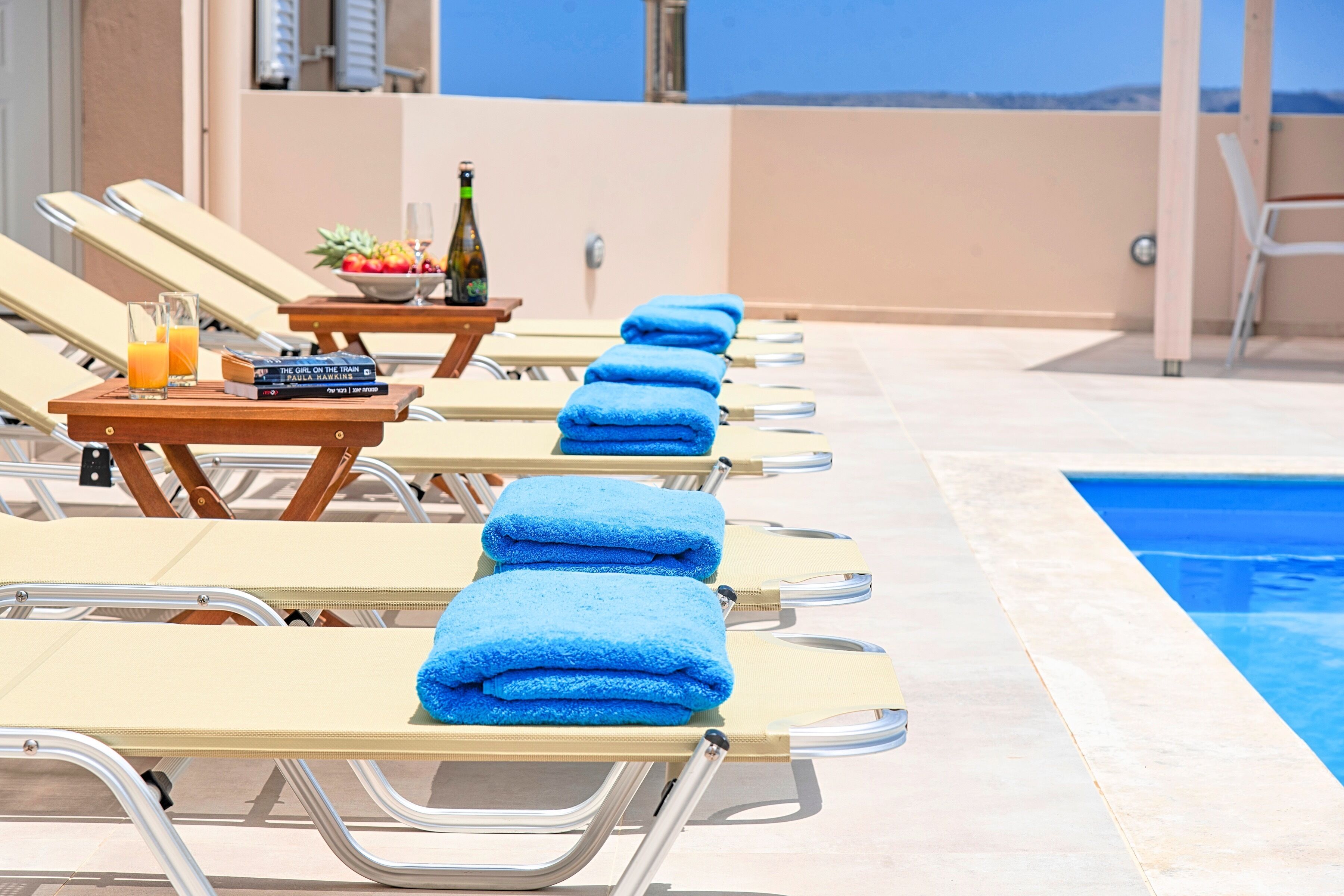 Ein Poolbereich mit Liegestühlen, Handtüchern, einem Tisch mit Flasche und Obst sowie Blick auf das Meer.