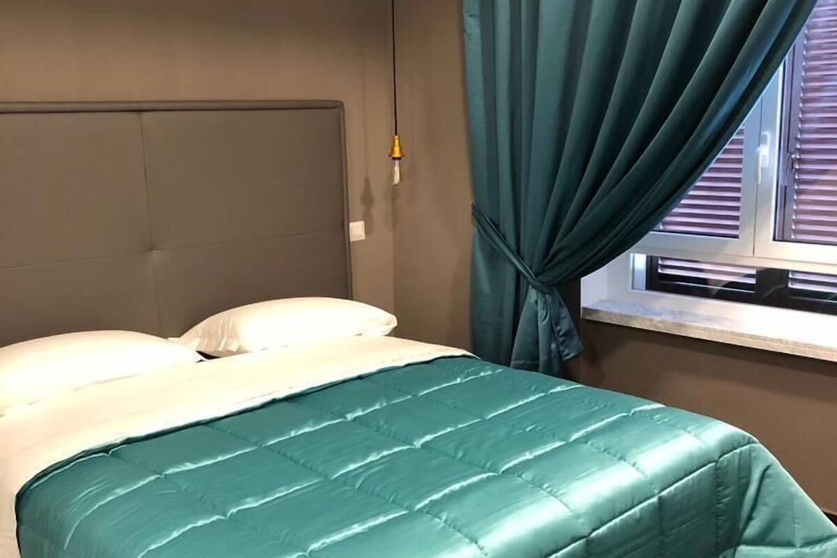 Una cama bien hecha con una colcha color turquesa, un cabecero gris y una ventana con cortinas.