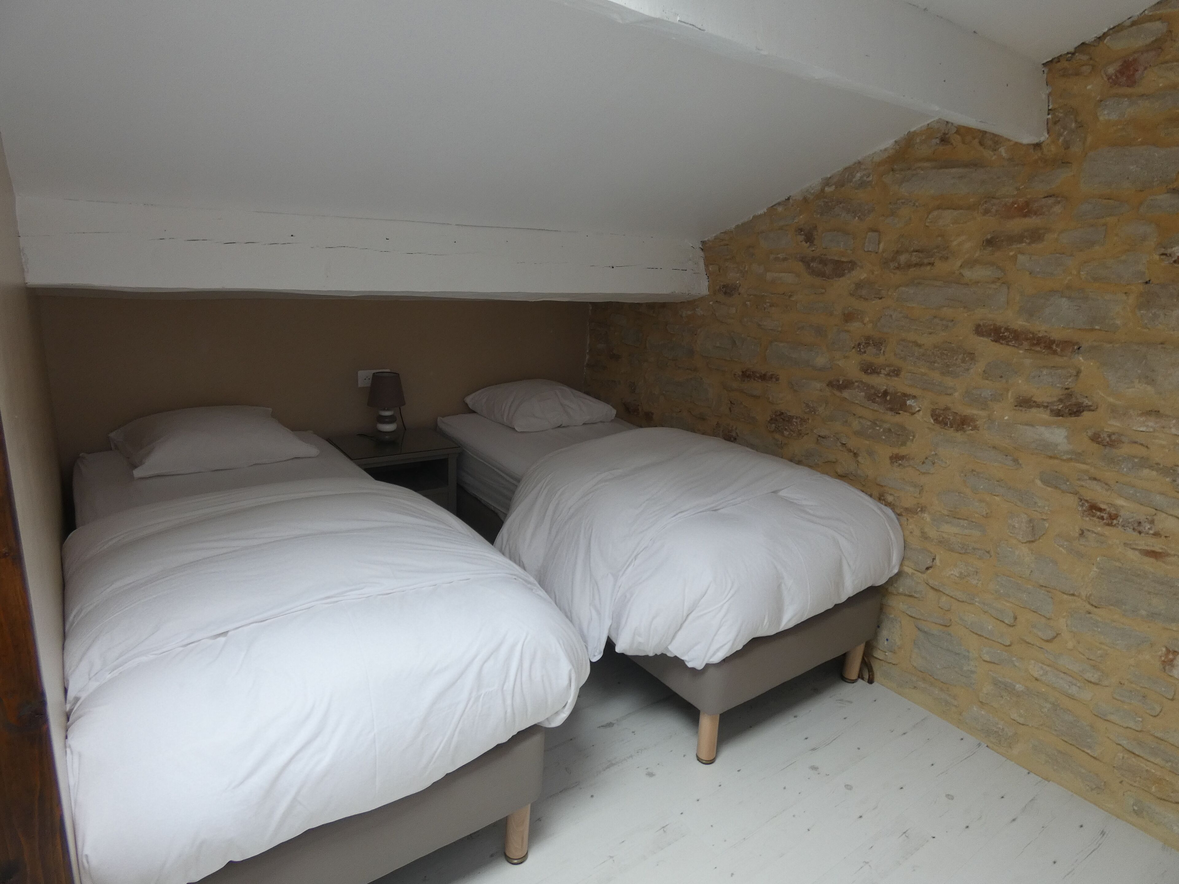 Une petite pièce à la lumière tamisée, avec deux lits simples, une table de chevet avec une lampe et un mur en pierre.