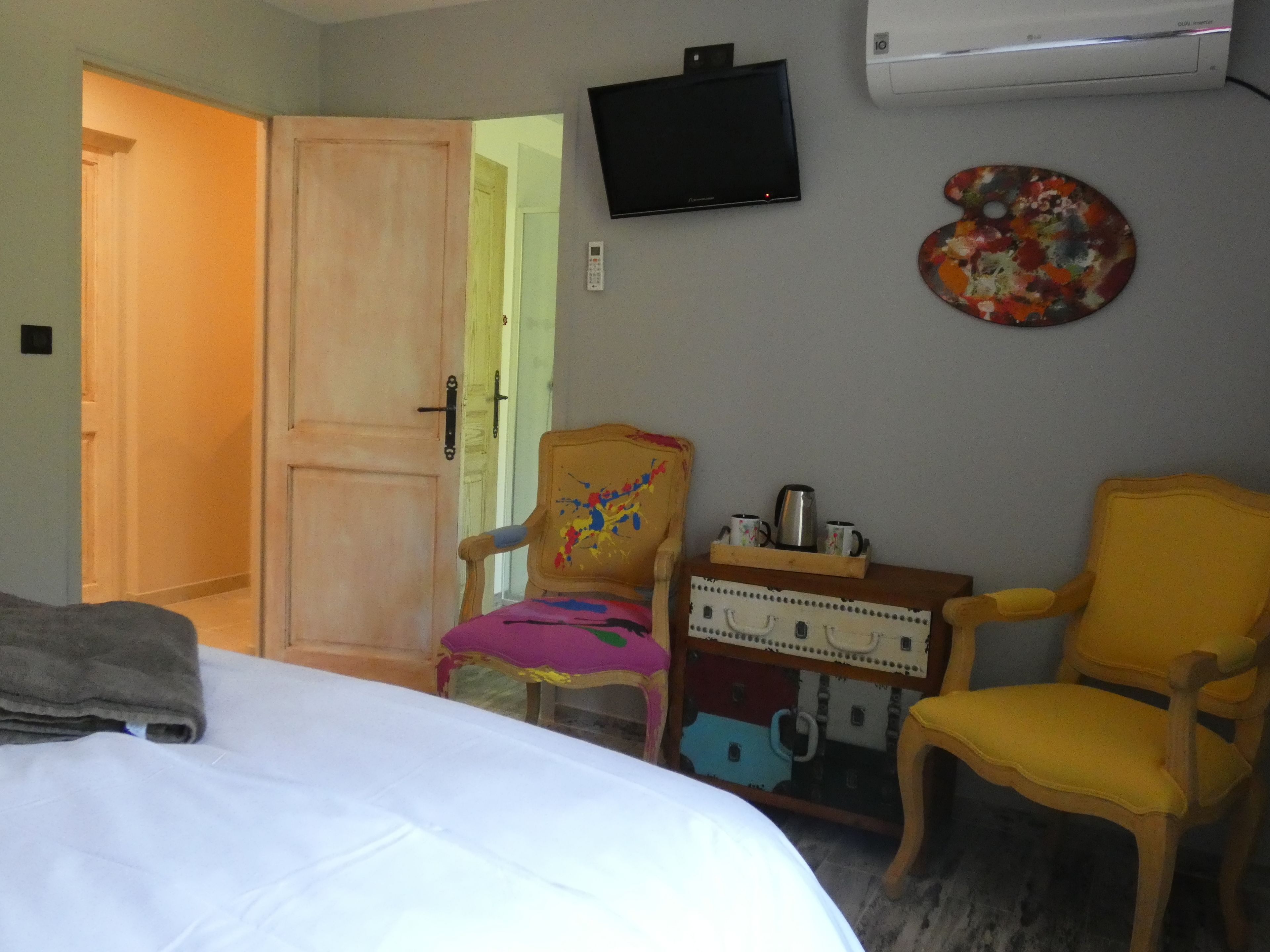 A hotel room with a bed, two chairs, a small table with a tray, a television, and an air conditioning unit.