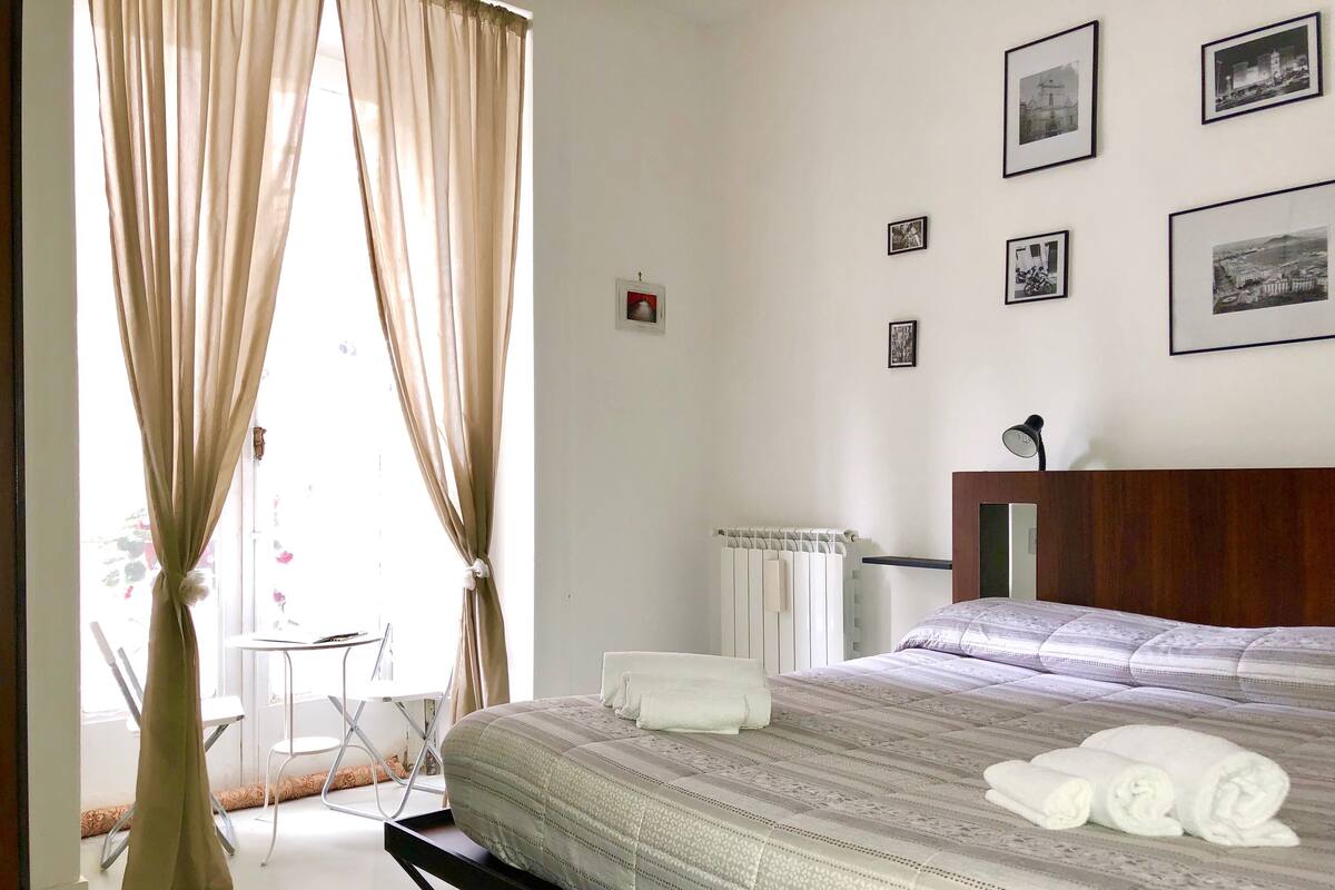Une chambre d’hôtel bien rangée, avec un lit, une chaise, un radiateur et un mur orné de tableaux encadrés.