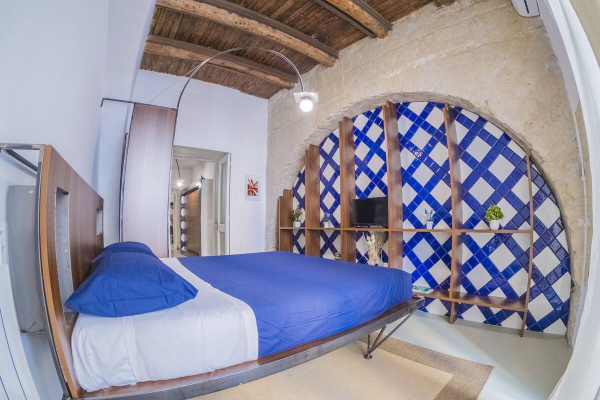 Un lit simple avec une couvre-lit bleue et blanche, dans une pièce dont on peut voir les poutres apparentes et qui possède un mur recouvert de carreaux.