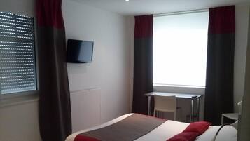 A hotel room with a bed, a TV mounted on the wall, a window with curtains, and a small table with a chair.