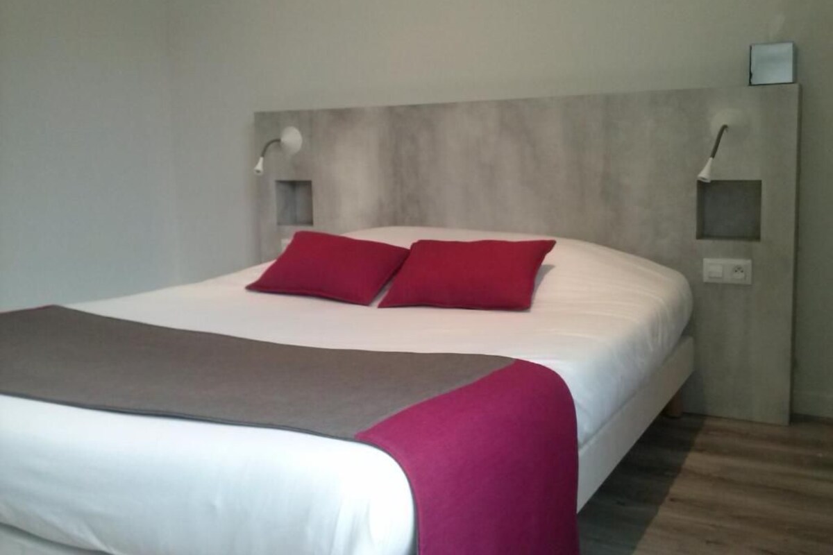 Una cama con ropa de cama blanca y dos almohadas rojas, un cabecero gris y una mesita de noche con una lámpara.