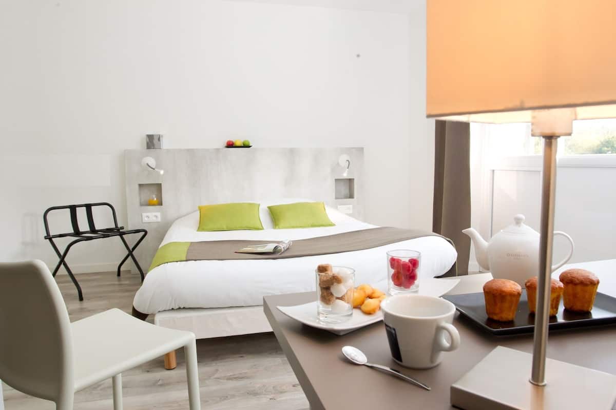 A modern hotel room with a bed, a dining table with breakfast items, a chair, and a lamp.