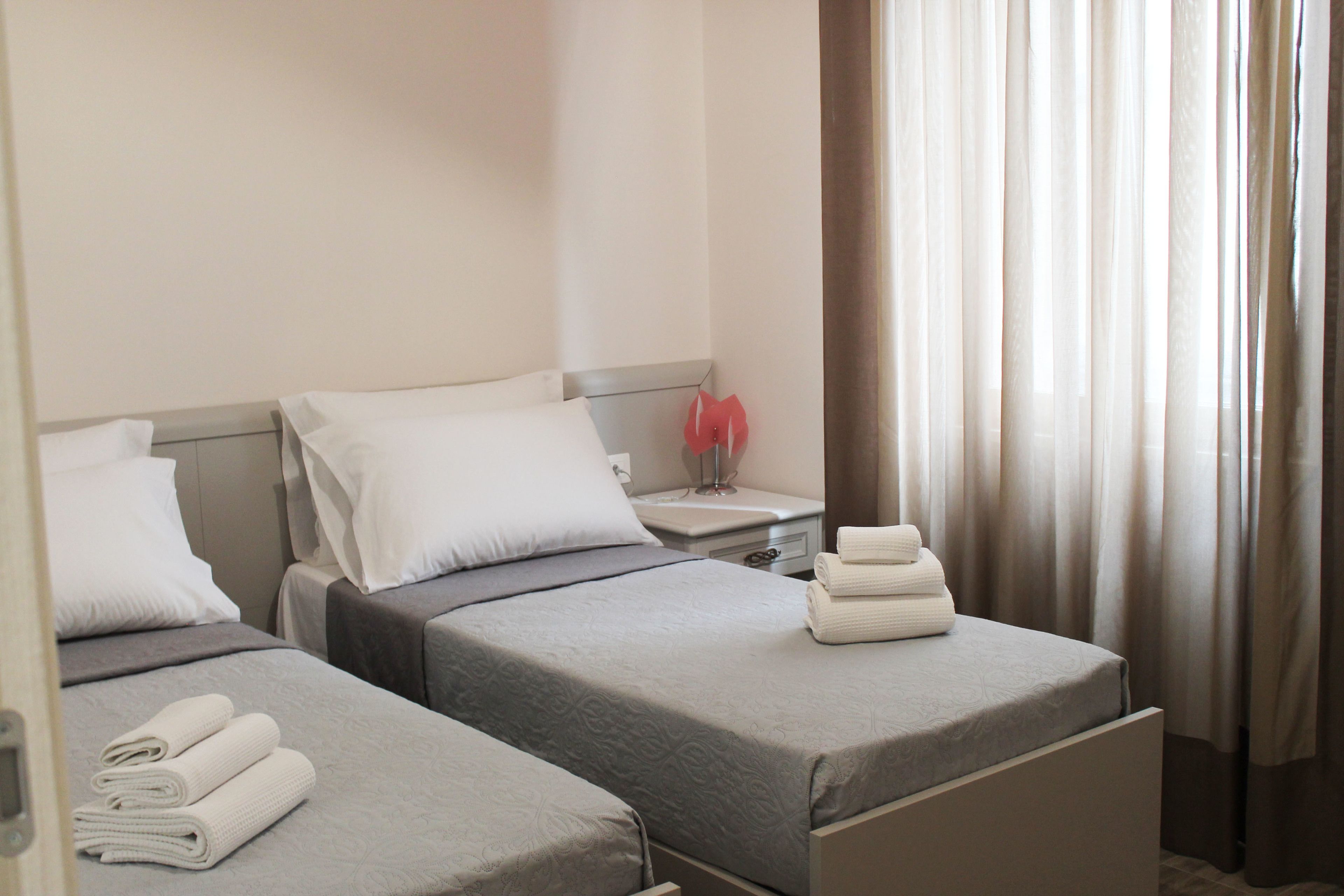 Habitación de hotel con dos camas, cada una con una almohada y una toalla doblada. Se aprecia una mesita de noche con una lámpara y un pequeño adorno.
