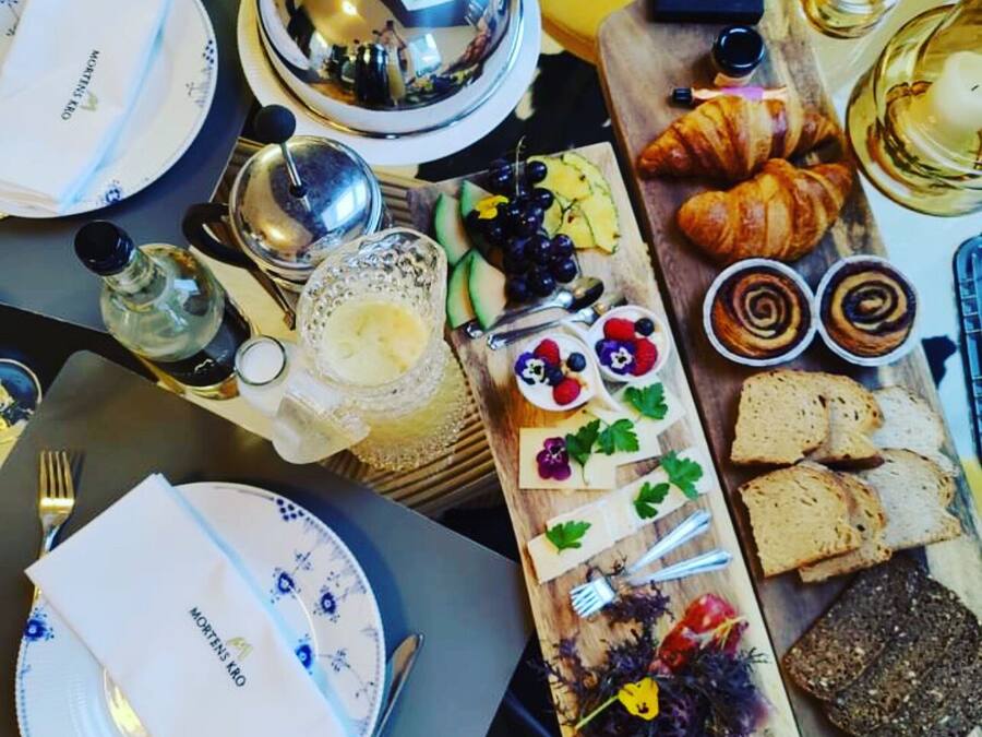 Una mesa lista para una comida, con platos, cubiertos, una botella, una copa y una tabla de madera con pan, pasteles y fruta.