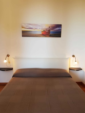 A bedroom with a bed, two bedside tables, and a painting of a boat on a beach.