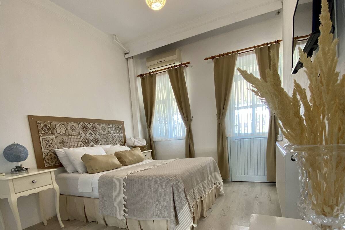Un dormitorio con cama, mesita de noche, cortinas y un jarrón con plantas secas.