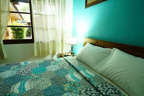 A bedroom with a bed, a nightstand, a lamp, and a window with curtains. - Mangga Bungalow (Denpasar)
