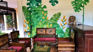 A room with a large tropical leaf mural, wooden furniture, and a small wooden cabinet. - Mangga Bungalow (Denpasar)