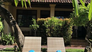 Two wicker lounge chairs with blue towels, positioned in front of a building with a tiled roof and a well-maintained garden. - Mangga Bungalow (Denpasar)