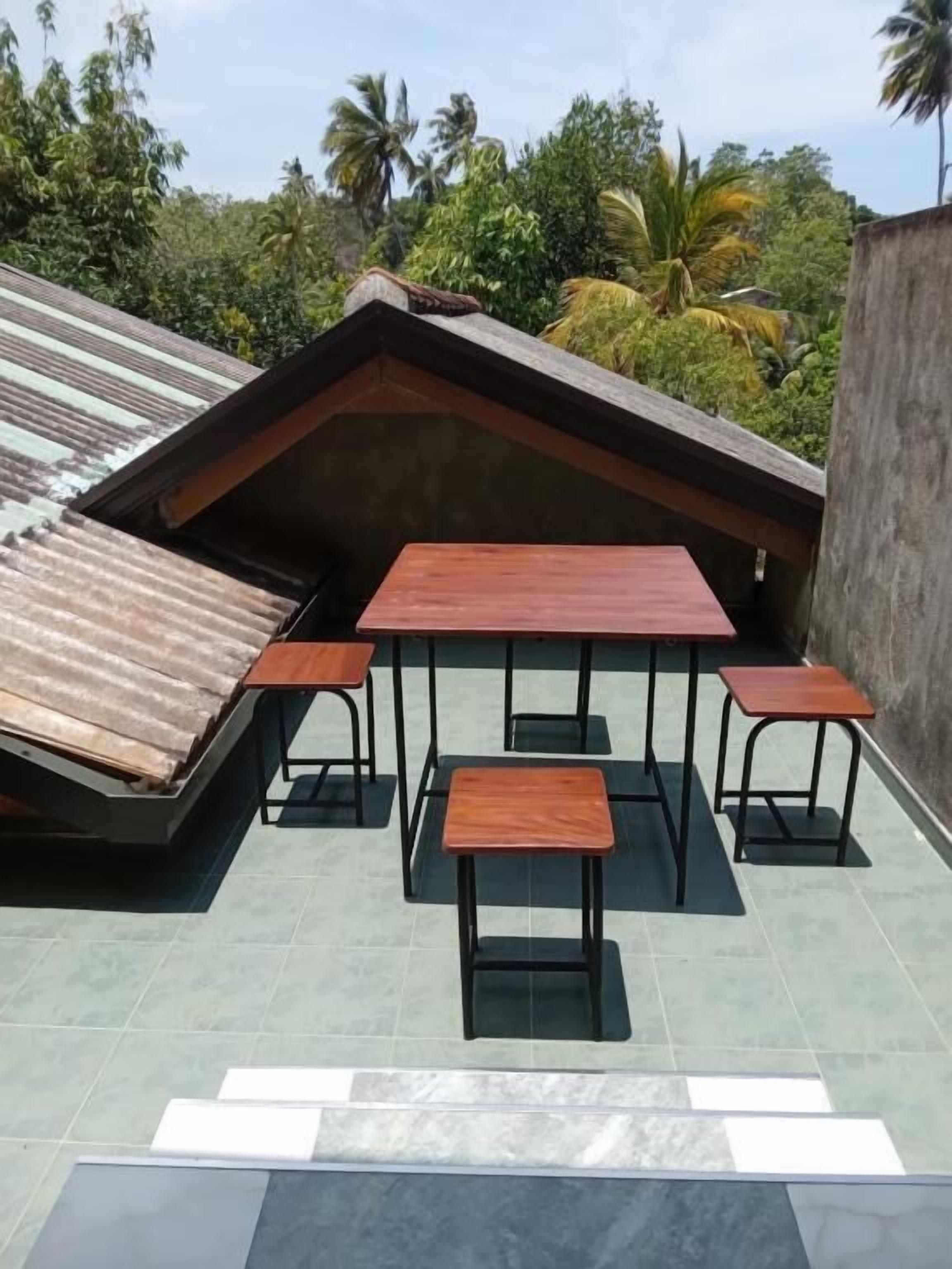 A rooftop terrace with a wooden table and benches, surrounded by a low wall and a view of tropical vegetation.