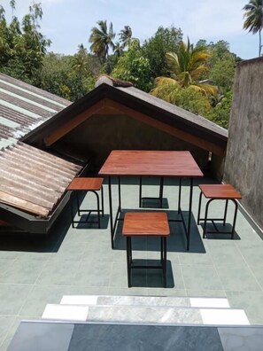 A rooftop terrace with a wooden table and benches, surrounded by a low wall and a view of tropical vegetation.