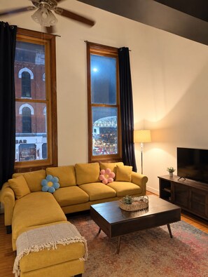 A living room with a sofa, a coffee table, a TV, and a lamp. There are two windows with curtains, and a ceiling fan is visible. - Steps from Broadway - The Lofts @ 107 - Unit 7, Steps from Broadway - The Lofts @ 107 - Unit 7 (Nashville)