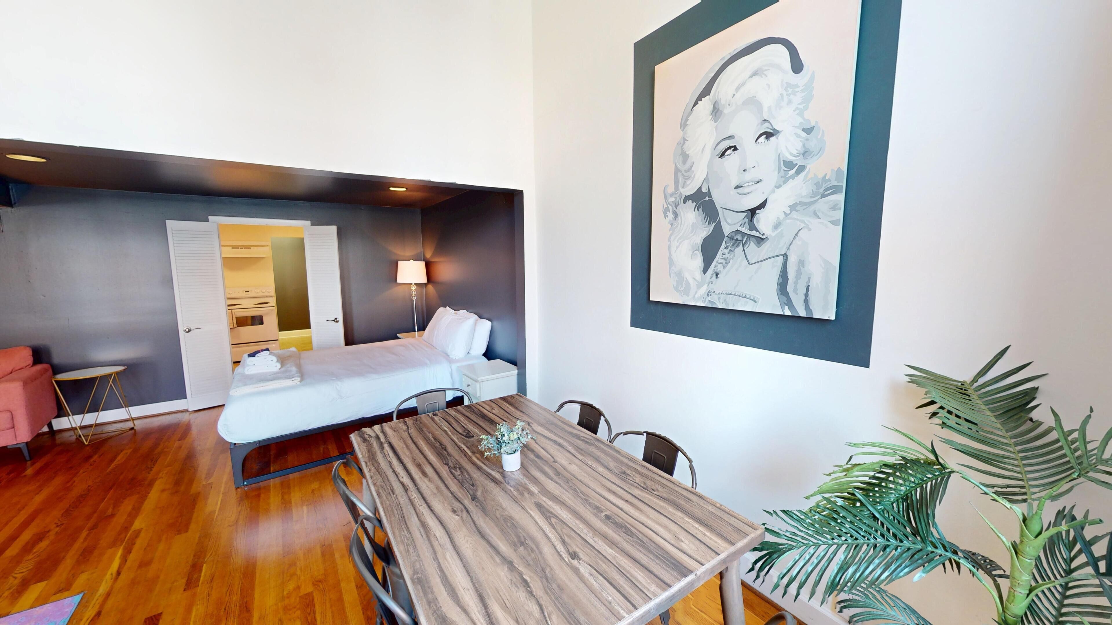 Una habitación de hotel moderna con suelo de madera, una cama con ropa blanca, una mesa de comedor, una pequeña planta en maceta y una obra de arte grande enmarcada en la pared.