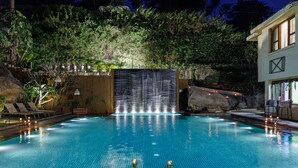 Una piscina con una cascada, rodeada de sillas de descanso y un edificio con balcón.