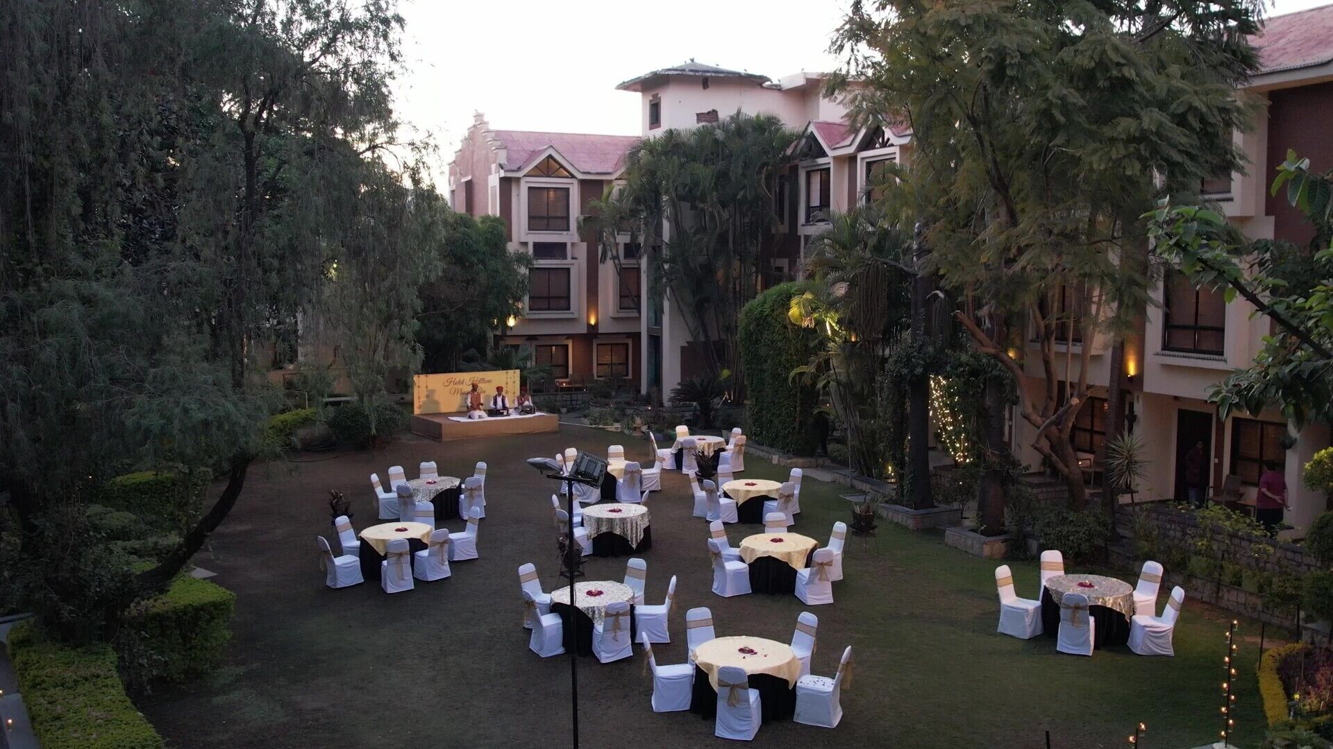 An outdoor event space with round tables and chairs, surrounded by lush greenery and a building in the background.