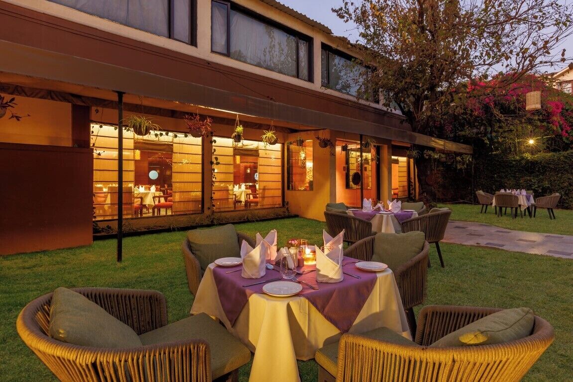 Un área de comedor al aire libre con mesas dispuestas para una comida, rodeada de sillas de mimbre y un edificio con grandes ventanales.