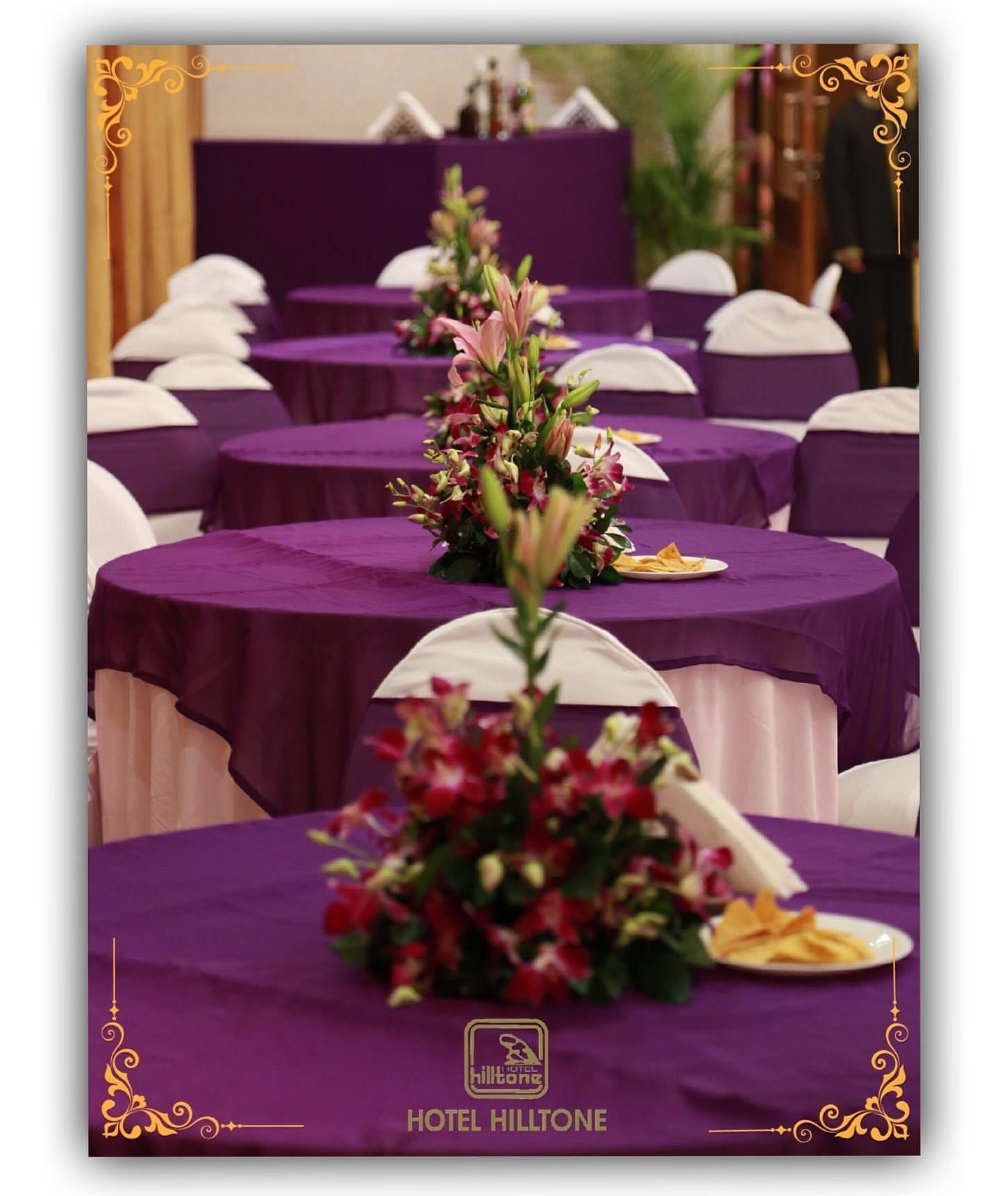 A hotel banquet hall with round tables covered in purple tablecloths, each with a floral centerpiece and white napkins.