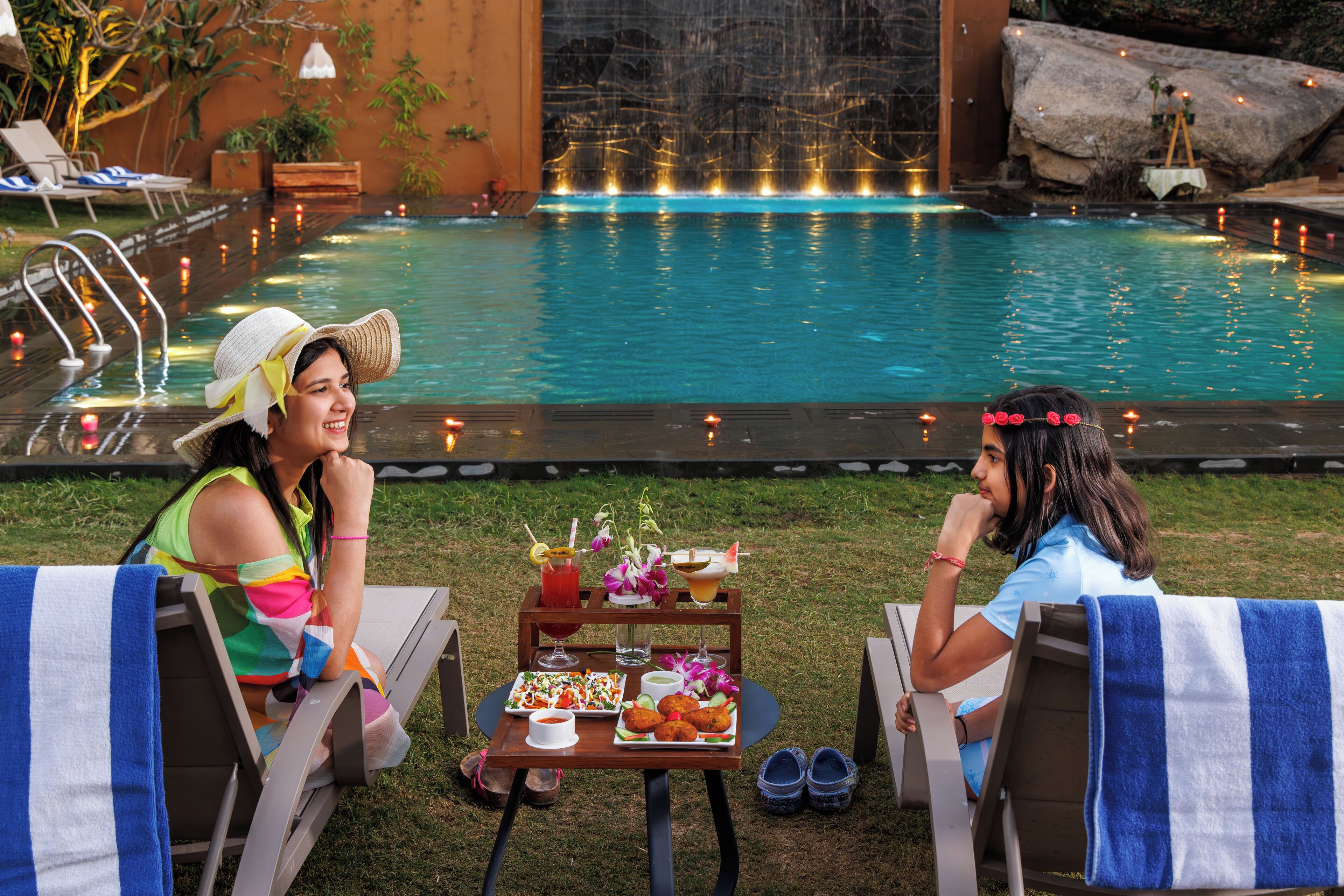 A poolside dining area with a table set for two, a pool with water features, and lounge chairs.