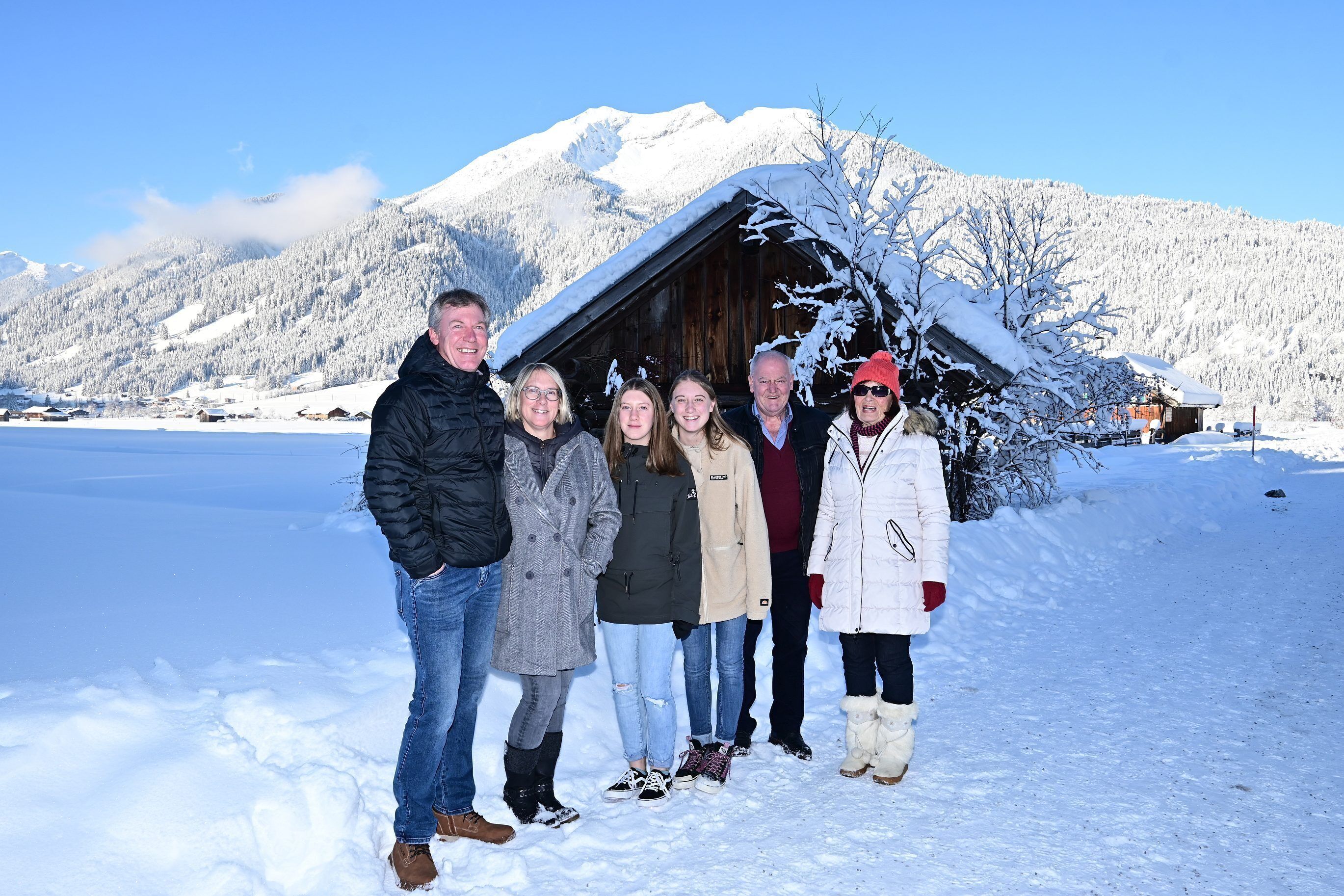 A group of people standing in a snowy landscape with a wooden cabin and mountains in the background.
