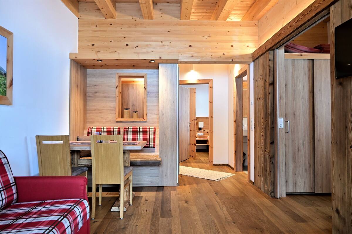 Une pièce chaleureuse avec un plafond en bois, un canapé à carreaux rouges, une table à manger en bois avec des chaises, et un coin cuisine visible à travers une porte ouverte.