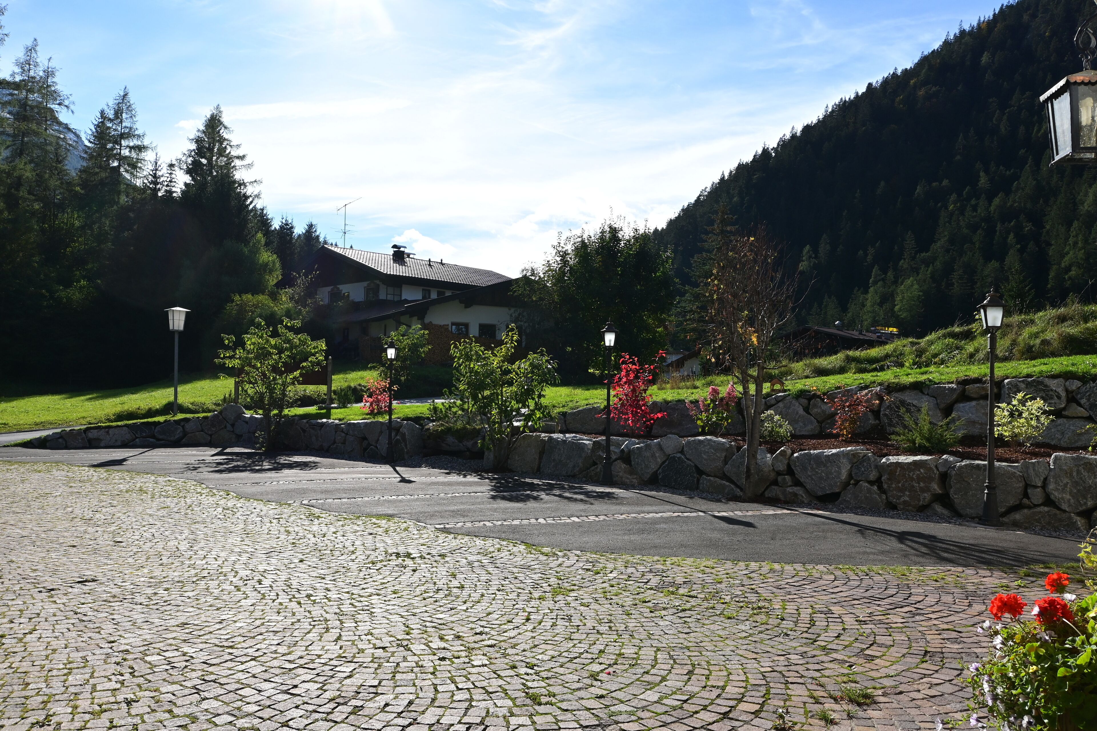 Un patio empedrado con un muro de piedra, un edificio con techo inclinado y un paisaje montañoso cubierto de bosque al fondo.