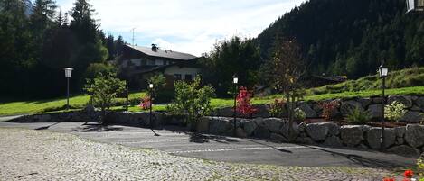 Un patio empedrado con un muro de piedra, un edificio con techo inclinado y un paisaje montañoso cubierto de bosque al fondo.